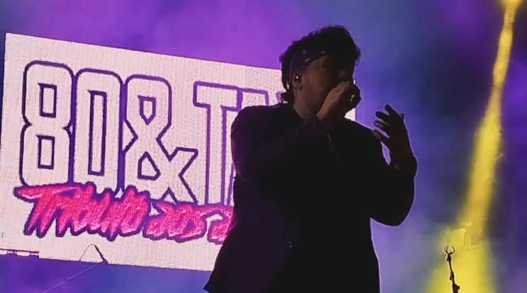 Vocalista dos 80&Tais em palco com o logotipo da banda ao fundo.